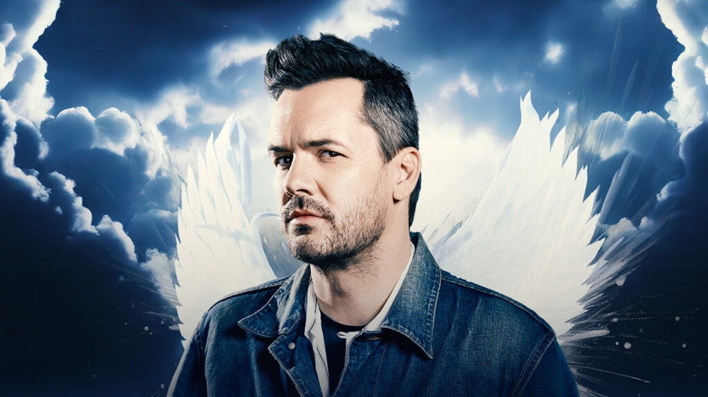 Comedian Jim Jefferies with angel wings posing against a clouded background.