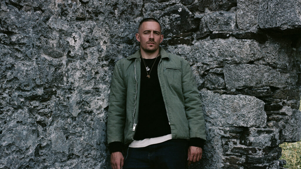 Dermot Kennedy poses in front of a rugged stone wall.